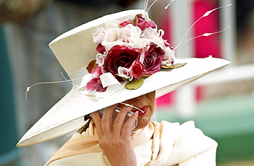 Race-goer wearing a cream hat trimmed with flowers at Royal Ascot Races while chatting on her mobile cell phone.