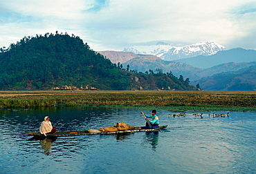 Man rowing a boat carrying a woman and some cargo in the foothills of the Himalayas, Nepal.  Behind is the Annapurna range of mountains.