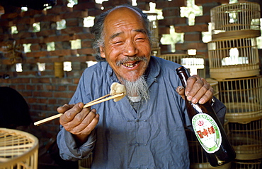 Market trader selling bird cages stops for a beer and his lunch using chopsticksduring a break in Peking (Beijing) China