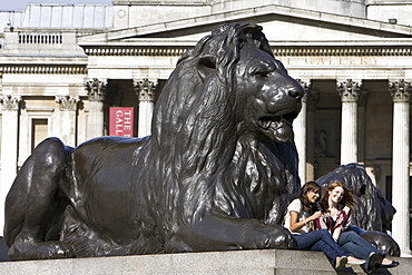 Tourists sit on lion statues in front of National Gallery in Trafalgar Square, London, United Kingdom