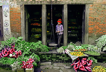 A young boy in a checked shirt and red knitted hat  in the doorway of a fruit and vegetable shop in Patan, Nepal