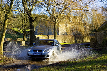 BMW 5 Series car drives through country lane ford, Swinbrook, Oxfordshire, United Kingdom