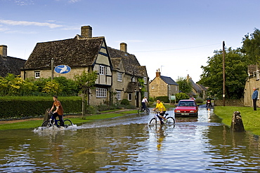 Children ride their bikes through  flood water in Minster Lovell, The Cotswolds, Oxfordshire, England, United Kingdom