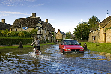 Car and boy on a bike try to move through flood water in Minster Lovell, Oxfordshire, England, United Kingdom