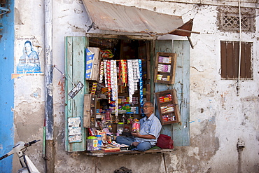 Stallholder selling sweets, tobacco and foods sits in shop window in old town in Udaipur, Rajasthan, Western India