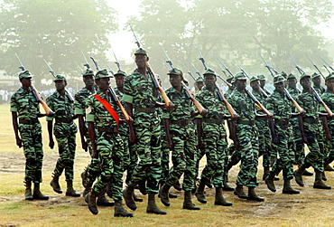 Soldiers carrying rifles on their shoulders marching in an Independence Day parade in Banjul, Gambia.  One man stands head and shoulders above the rest. He is likely to be more than seven feet tall.