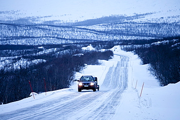 Car with trailer towing a snowmobile travels through arctic wilderness at nightfall by Kilpisjarvi on route from Norway into Finland