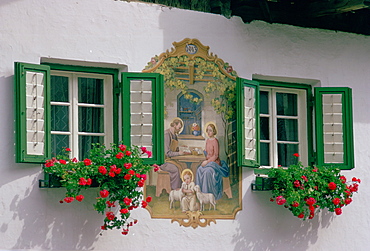 In the Austrian Tyrol a farmhouse mural depicting baby Jesus with a halo hugging lambs with his parents Mary and Joseph nearby.This religious mural  painted in 1943  is sited between two flower troughs planted with geranium plants below windows with shutters.