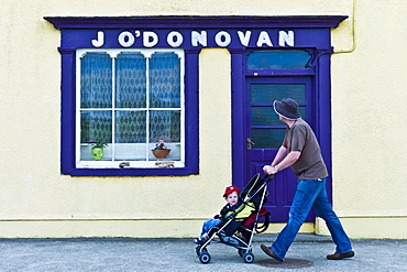 Man wheels child in stroller past J'ODonovan shop front at Courtmacsherry, County Cork, Ireland