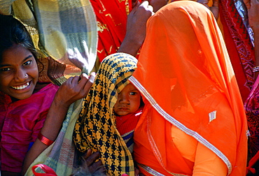 A  veiled woman, in brightly coloured robes holding her baby while watching a village festival in Nalu, Rajasthan, India