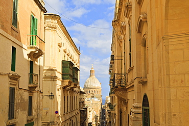 View down a narrow street with golden stone, green shutters and balconies, to the Carmelite Church and its dome, Valletta, Malta, Europe 