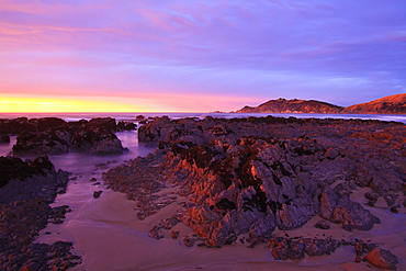 Sunrise casts a red pink hue on rocks of a beach looking towards Nugget Point, South Island, New Zealand, Pacific 