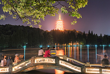 Leifeng Pagoda with visitors sitting on railings of an illuminated stone arch bridge at West Lake, Hangzhou, Zhejiang, China, Asia