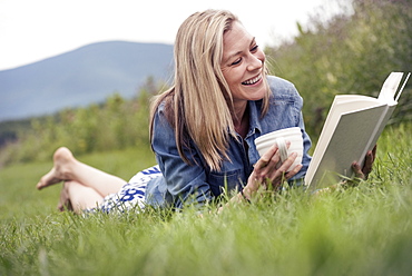 A woman lying on the grass holding a tea cup and reading a book.