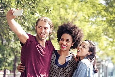 Three people, a man and two women, taking selfies in a city park