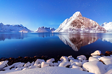 The snowy peaks are reflected in the frozen sea on a starry winter night, Reine Bay, Nordland, Lofoten Islands, Arctic, Norway, Scandinavia, Europe