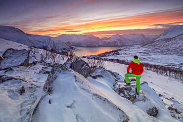 Hiker admires the frozen sea surrounded by snow framed by the orange sky at sunset, Torsken, Senja, Troms County, Arctic, Norway, Scandinavia, Europe