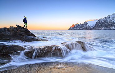 Hiker admires the waves of the icy sea crashing on the rocky cliffs at dawn, Tungeneset, Senja, Troms county, Arctic, Norway, Scandinavia, Europe