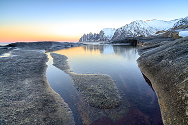 Orange sky at sunset reflected on snowy peaks and the frozen sea surrounded by rocks Tungeneset, Senja, Troms County, Arctic, Norway, Scandinavia, Europe