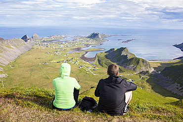 Couple on top of peak admire sea framing the village of Sorland, Vaeroy Island, Nordland county, Lofoten archipelago, Norway, Scandinavia, Europe