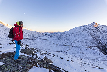 Hiker on top of the rocky crest admires the snowy peaks of Fjordbotn, Lysnes, Senja, Troms, Norway, Scandinavia, Europe