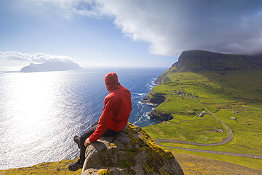 Hiker sitting on rocks looking at the ocean, Gasadalur, Vagar Island, Faroe Islands, Denmark, Europe