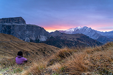 Rear view of woman sitting on grass admiring sunrise on Marmolada and Sass Pordoi, Passo Sella, Dolomites, South Tyrol, Italy, Europe