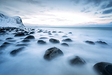 Rocks in the cold sea and snow capped mountains under the blue light of dusk, Unstad, Lofoten Islands, Arctic, Northern Norway, Scandinavia, Europe