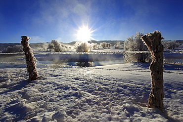 Barb wire fence in winter landscape with hoarfrost, steeming river, brilliant sunshine and blue sky, utah, usa