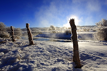 Barb wire fence in winter landscape with hoarfrost, steeming river, brilliant sunshine and blue sky, utah, usa