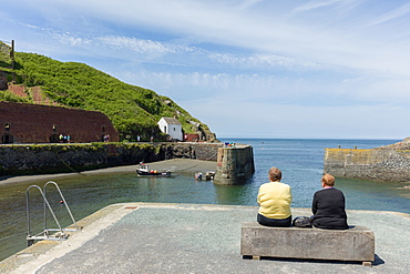 Two ladies on a bench at Porthgain harbour as the Pembrokeshire coast path extends up the cliffs beyond, Wales, United Kingdom, Europe