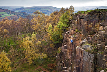 A rock climber ascends a cliff face formed by historic quarrying at Bole hill quarry on an autumn day in the Peak District, Derbyshire, England, United Kingdom, Europe