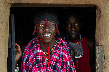 Two Maasai tribesmen in costume wearing traditional dress stood in doorway of mud house, Maasai Mara, Kenya Africa