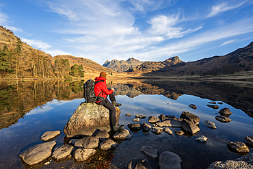 A walker wearing red jacket and backpack sat admiring the mirror conditions at Blea Tarn, Lake District, Cumbria