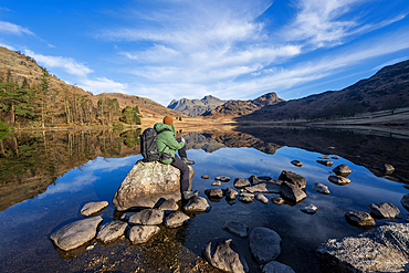 A walker with backpack sat admiring the mirror conditions at Blea Tarn, Lake District, Cumbria