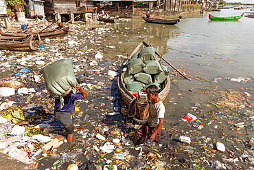 Two men unloading sacks of produce from a small boat in Sittwe harbour, wading through polluted water full of plastic rubbish, Sittwe, Rakhine, Myanmar (Burma), Asia