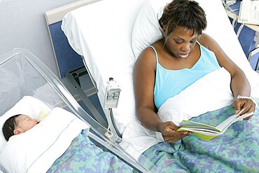 Mother reading magazine in bed of maternity hospital, her newborn baby beside