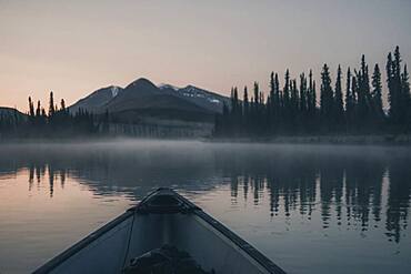 A Canoe glides over the quiet water of a river while the sky starts to glow softly, shortly before the sunrise. Yukon Territory, Canada