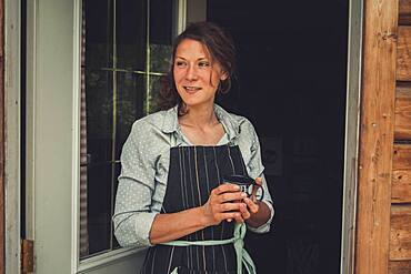 Thirty year old attractive brunette woman standing in the doorframe with a cup of coffee in her hands. Yukon Territory, Canada