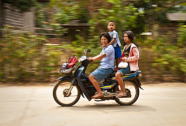 Family riding motorcycle through village along the Mae Taeng River in rural Chiang Mai Province, Thailand.