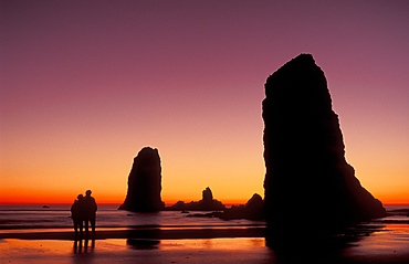 Haystack Rock & sea stacks at sunset with couple on beach; Cannon Beach, Oregon coast.