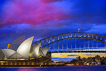 World famous Sydney Opera House and Harbour bridge at sunset. Blurred clouds and lights of landmarks reflect in blurred waters of Harbour. Sydney, New South Wales, Australia