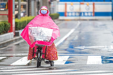 Shanghai, China, 25th Jan 2020, A woman wearing a mask uses rain poncho as she bikes on road