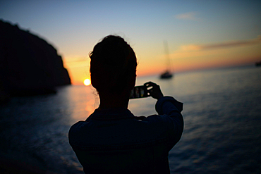 Silhouette of a young woman taking a photo of the sunset and a sail boat on the beach