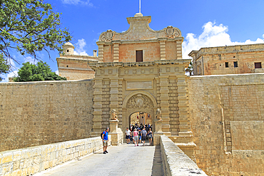 Tourists walking over moat bridge through entrance gateway of medieval city of Mdina, Malta, Mediterranean, Europe