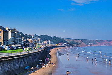 Sunny beach day with people enjoying the seaside, clear blue sky, and coastal buildings in the background in Filey, England.