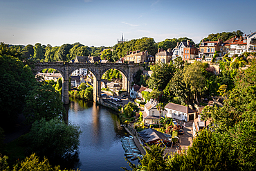 Scenic view of an old arched bridge over a tranquil River Nidd with surrounding greenery and houses in a quaint village in Knaresborough, North Yorkshire, England, United Kingdom, Europe