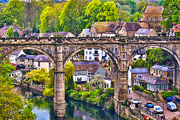 A picturesque view of a stone bridge arching over a river in a quaint village, surrounded by lush greenery and historic buildings.