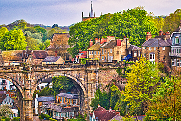 Scenic view of a historic town with a prominent stone bridge and lush greenery. Traditional architecture and a church spire add to the charm.