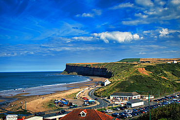 Sunny coastal landscape with cliff, beach, and winding road leading to a small seaside village in Saltburn-by-the-Sea, England.